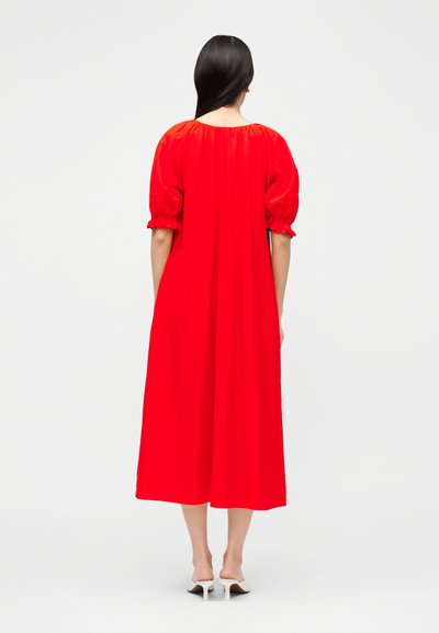 Γυναίκα με μακριά σκούρα μαλλιά, που φοράει ένα φωτεινό κόκκινο φόρεμα μέχρι τη μέση της γάμπας με φουσκωτά κοντά μανίκια και λευκές γόβες, γυρισμένη με την πλάτη σε απλό φόντο.