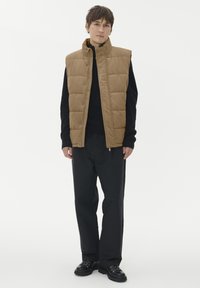 Tannfarbenes Steppweste mit gestepptem Design, hohem Kragen und Druckknöpfen vorne, getragen über einem schwarzen Pullover, kombiniert mit schwarzen Hosen und klobigen schwarzen Schuhen.