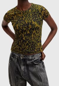 Person med en kortärmad topp i grön och svart leopardmönster och löst sittande, urblekta svarta jeans, med händerna vilande på höfterna.