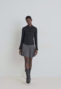 Schwarzes langärmeliges Hemd mit Kragen, kombiniert mit einem grauen Faltenrock. Das Model trägt schwarze Stiefel bis zum Knie. Glatter Stoff, minimalistisches Design.