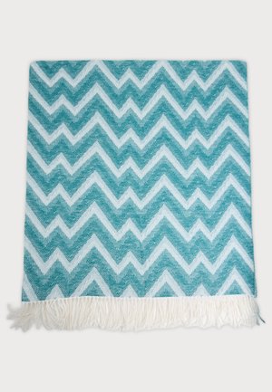 Serviette tissée turquoise et blanche, ornée d’un motif en zigzag répétitif et de franges blanches le long d’un bord, présentée à plat sur un fond clair.