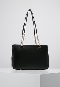 Sac à main rectangulaire noir avec chaîne dorée et doubles anses en cuir noir sur un piédestal blanc.