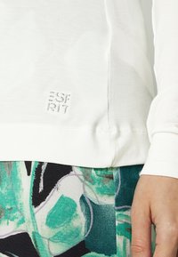 Biała koszula z długim rękawem z subtelnym haftowanym logo "Esprit" u dołu. W zestawie z kolorową, wzorzystą odzieżą w odcieniach zieleni i czerni.