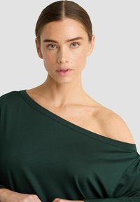 Groene, off-shoulder top van zacht materiaal. Heeft een ruime halslijn en lange mouwen, wat zorgt voor een ontspannen pasvorm. Vloeiende textuur en val.