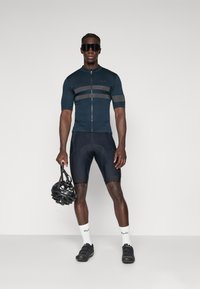 Maillot de cyclisme bleu foncé avec des rayures grises, à manches courtes et fermeture éclair sur le devant. Short noir et chaussures, tenant un casque noir. Chaussettes blanches.