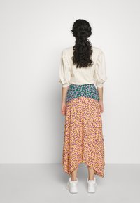 Vestido largo floral con un vibrante estampado en amarillo y púrpura, que presenta una cinturilla verde. Tejido ligero, dobladillo asimétrico. Zapatillas blancas.