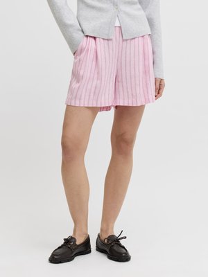 Persoon draagt lichtroze gestreepte korte broek, grijs vest met knopen en zwarte veterschoenen, staand met handen in de zakken.