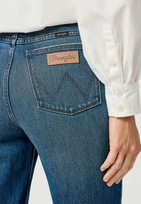 Jeans de mezclilla azul con un parche de cuero de Wrangler en el bolsillo trasero, que presenta un diseño de costura en zigzag. La tela tiene un acabado texturizado.