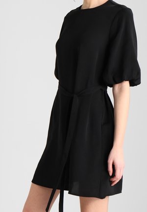 Femme portant une robe courte noire avec des manches bouffantes et une ceinture à la taille sur un fond clair uni.