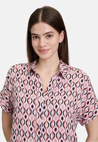 Camicia a maniche corte con bottoni, caratterizzata da un motivo astratto colorato in rosa, nero e verde su uno sfondo chiaro. Tessuto leggero.
