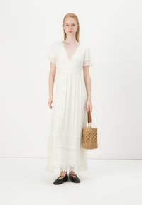 Vestido maxi branco com mangas curtas em estilo babado, detalhes em renda e um decote em V profundo. Combinado com uma bolsa de vime e sapatos pretos.