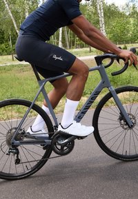 Bicicleta de carretera gris con un cuadro ligero, ruedas negras y componentes Shimano. Ciclista con camiseta oscura, pantalones cortos negros y zapatillas de ciclismo blancas.