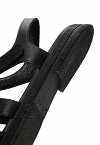 Sandale en cuir noir avec brides réglables et semelle en caoutchouc texturée, présentée en gros plan sous un angle mettant en valeur les détails de la semelle et des brides.