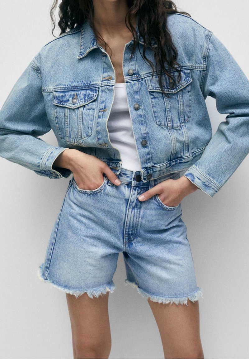 Ljusblå denimjacka med en kortad design, två bröstfickor och metallknappar, tillsammans med högmidjade denimshorts med fransiga kanter.
