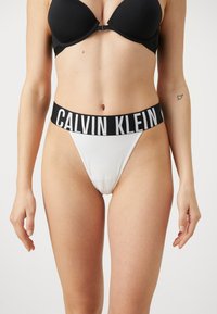 Calvin Klein witte thong met een brede zwarte tailleband waarop het logo "CALVIN KLEIN" staat. De stof is glad en rekbaar voor comfort.