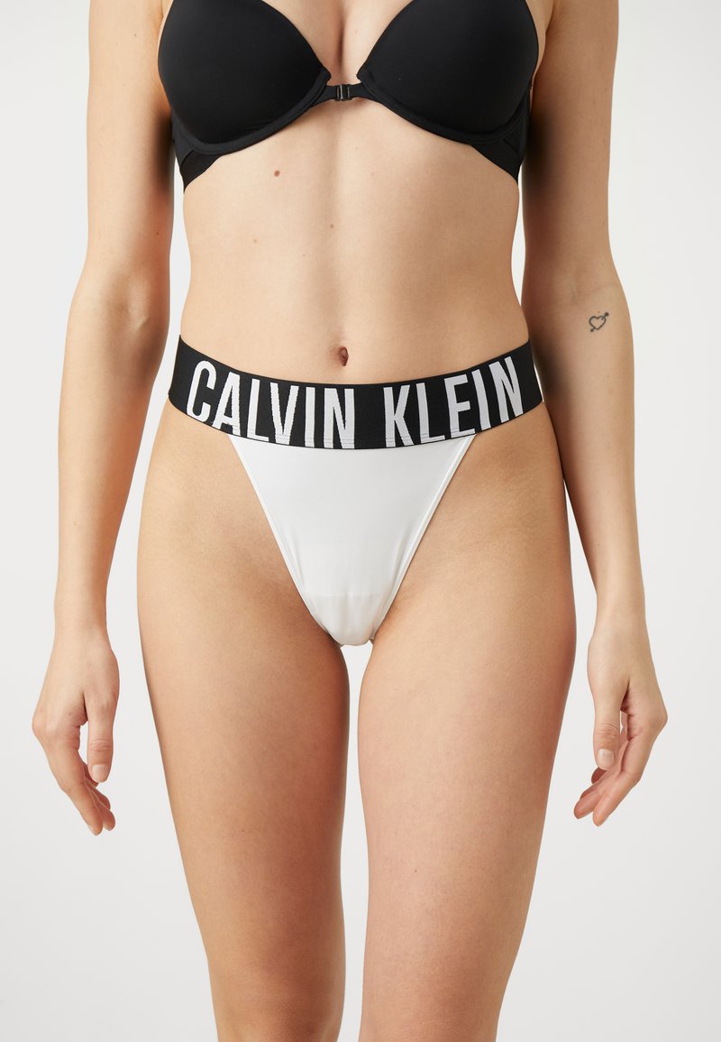 Calvin Klein witte thong met een brede zwarte tailleband waarop het logo "CALVIN KLEIN" staat. De stof is glad en rekbaar voor comfort.