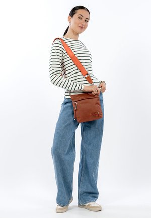 Sac bandoulière synthétique marron avec finition texturée, sangle orange réglable, fermeture éclair et détail du logo à l'avant. Le modèle porte un haut rayé décontracté et un jean.