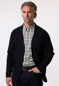 Námořnický svetr s texturovaným povrchem, s jednou náprsní kapsou, nošený přes světle kostkovanou košili v modré a krémové barvě, s tmavými džíny.