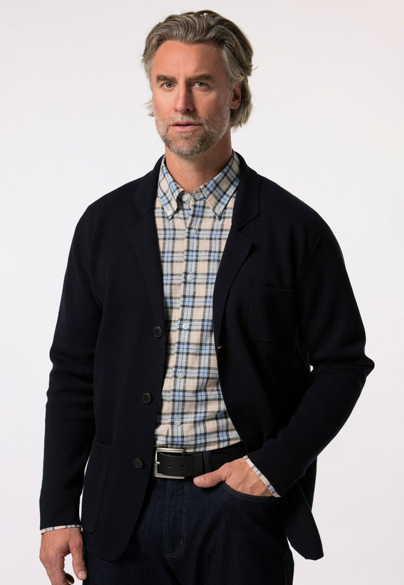 Námořnický svetr s texturovaným povrchem, s jednou náprsní kapsou, nošený přes světle kostkovanou košili v modré a krémové barvě, s tmavými džíny.