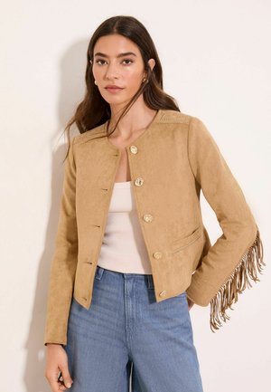 Woman wearing a tan suede jacket with long fringe on sleeves, gold buttons, white tank top, and blue high-waisted jeans against a light background.