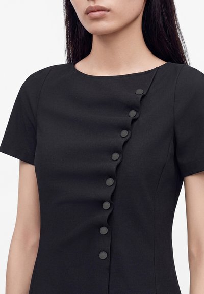 Femme portant une robe noire à manches courtes avec une rangée diagonale de boutons ronds noirs et une finition subtilement froncée sur l'encolure avant.