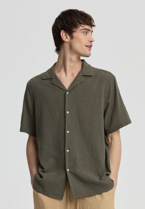 Giovane uomo con una camicia verde oliva a maniche corte abbottonata e pantaloni beige, che guarda di lato con le mani in tasca.