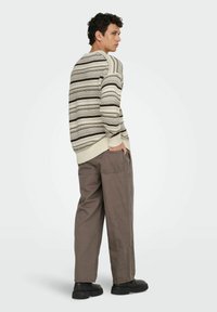 Sweter w paski w odcieniach beżu, brązu i kremu, z dzianinową teksturą; zestawiony z luźnymi spodniami w odcieniu szaro-brązowym; czarne buty. Ukazany widok boczny.