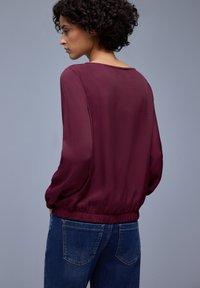 Blouse bordeaux avec taille élastique, manches longues en voile transparent et col rond, associée à un jean en denim bleu. Vue de dos montrée.
