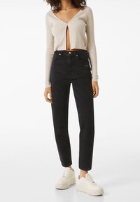 Jeans de tiro alto negros combinados con un cárdigan corto de canalé beige. La modelo lleva zapatillas de plataforma blancas. Diseño simple y estilo ajustado.