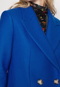 Cappotto blu a doppio petto con una trama testurizzata, rever affilati e bottoni in tonalità argento, indossato sopra un dolcevita nero a fiori.