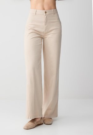 Beżowe, szerokie spodnie z wysokim stanem, noszone z dopasowanymi beżowymi mokasynami, skrócone w talii, na tle jednolitego jasnego tła.