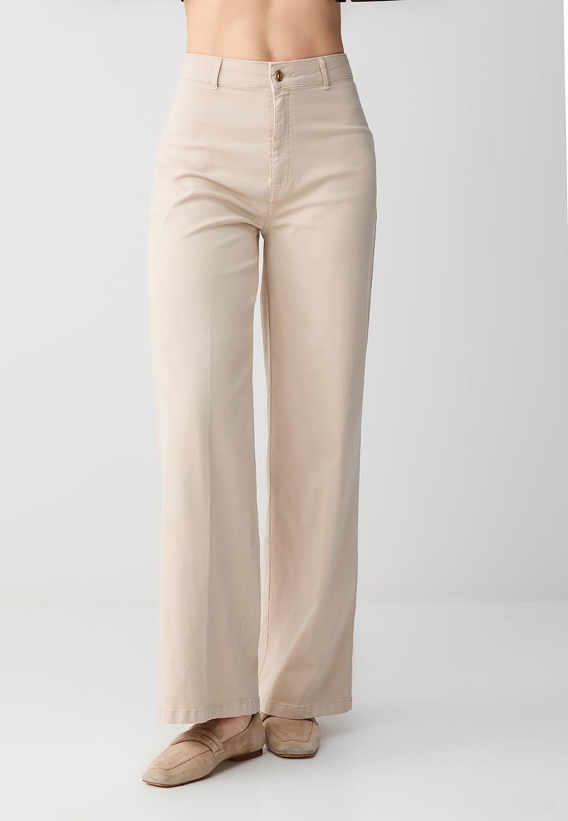 Beżowe, szerokie spodnie z wysokim stanem, noszone z dopasowanymi beżowymi mokasynami, skrócone w talii, na tle jednolitego jasnego tła.