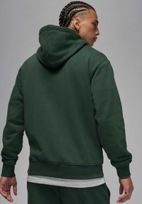 Zielona bluza z kapturem wykonana z miękkiego materiału, z długimi rękawami, kieszenią kangurka oraz ściągaczami na mankietach i u dołu. Widok od tyłu.