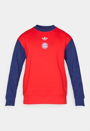 Červená mikina s tmavomodrými rukávmi. Má okrúhly výstrih, rebrované manžety a logo Adidas nad kruhovým emblémom Bayern Mníchov.