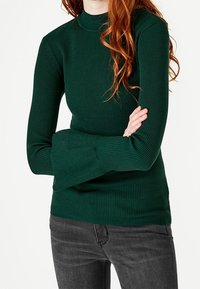 Haut vert côtelé à manches longues avec col montant et poignets évasés, associé à un jean noir ajusté.