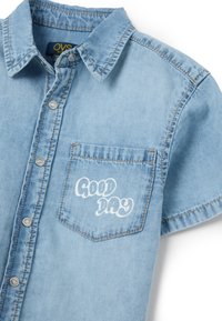 Denim overhemd in lichtblauw, met korte mouwen, met knopen en een kraag. Heeft een borstzak met de tekst "GOEDE DAG" in het wit.
