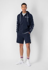 Felpa con zip nella tonalità navy, con dettagli del logo ai lati, abbinata a pantaloni corti coordinati. Presenta lacci bianchi e calze a contrasto con strisce rosse e blu.