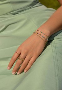 Pulseras de cuentas multicolores y anillos dorados en capas adornan una mano que descansa sobre un fondo de tela verde menta. Las uñas suaves presentan acentos florales.