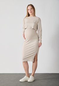 Completo materno a coste in beige, composto da un top corto a maniche lunghe e una gonna di lunghezza media con uno spacco laterale, abbinato a delle sneaker bianche.