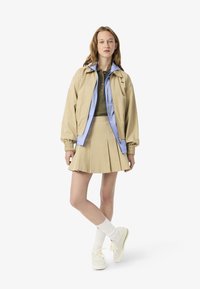 Giubbotto bomber beige chiaro con una fodera interna blu, abbinato a un polo verde e una gonna plissettata beige. Calze bianche e scarpe chiare completano il look.
