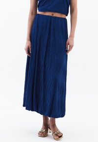 Blauwe geplooide maxi rok met een zachte textuur en elastische tailleband. Gecombineerd met metallic sandaaltjes met bandjes. Eenvoudige achtergrond.