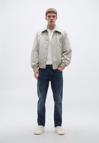 Svjetlo bež bomber jakna sa zaobljenim ovratnikom, zatvaranjem na gumbe i vezanim detaljem; uparena s tamnoplavim trapericama i svijetlim tenisicama.