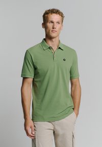 Groene poloshirt met een kraag, voorzien van drie knopen en een klein hexagonaal logo op de linkerborst. Korte mouwen, gladde textuur.