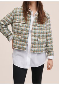 Femme portant une veste en tweed multicolore avec poches avant sur une chemise blanche boutonnée et un pantalon noir, debout devant un fond neutre.