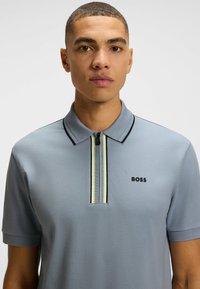 Jasnoniebieska koszulka polo z zamkiem błyskawicznym przy kołnierzyku, ozdobiona czarnymi detalami i pionowym wzorem w paski. Logo umieszczone jest po lewej stronie na klatce piersiowej.