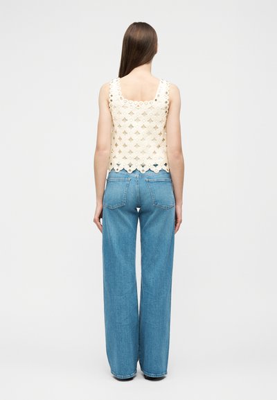 Femme de dos portant un haut en crochet crème sans manches et un jean à taille haute, jambes larges, bleu, sur un fond clair uni.