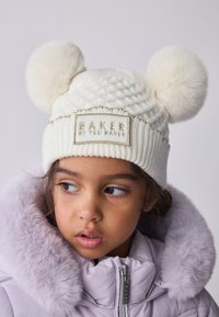 Fehér kötött sapka két bolyhos pomponnal és egy "BAKER by TED BAKER" feliratú varrt címkével. Halványlila, szőrme galléros dzsekivel viselve.