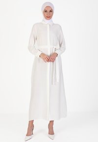 Long white dress with a button-down front, collar, and tied belt at the waist. Features long sleeves and a straight cut, made from lightweight fabric.