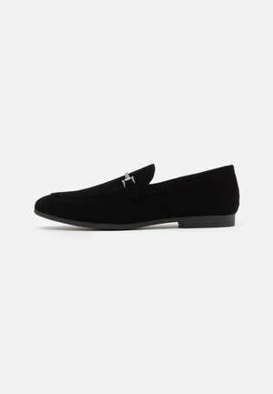 Black velvet loafers with a sleek design, featuring a silver-tone accent and a rounded toe. The sole is flat and textured for grip.