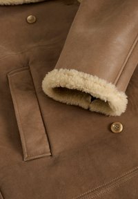 Hnědá semišová bunda s měkkou béžovou beranicí v límci a na manžetách, s knoflíkovým zapínáním a přední kapsou.